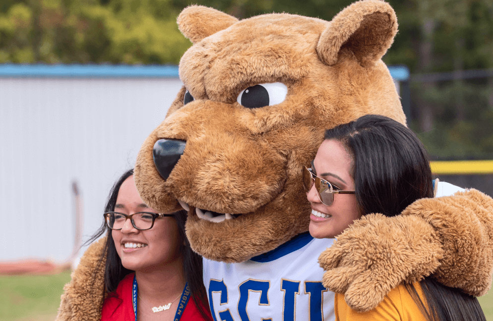 GCU mascot Roary the lion with his arms around two female students, smiling at a camera