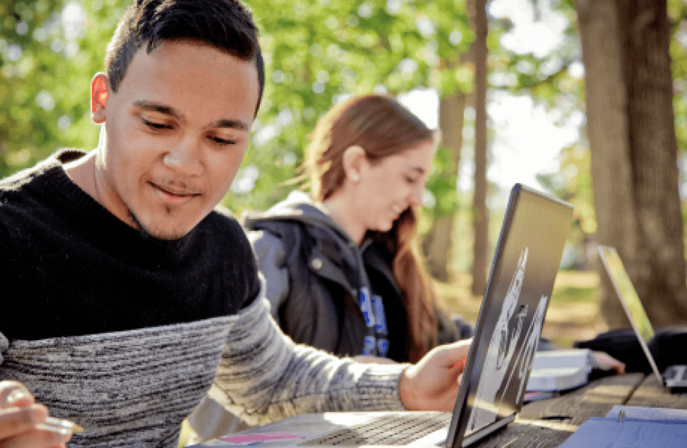 Two students studying outside on a picnic table with laptops