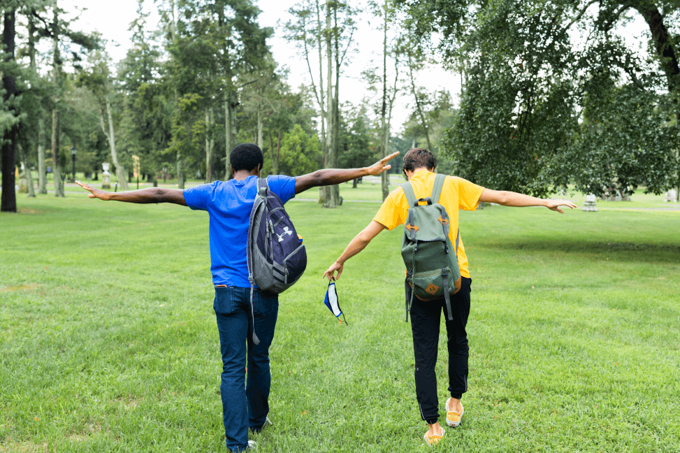 Two male students walking away from camera into a grassy field with their arms spread